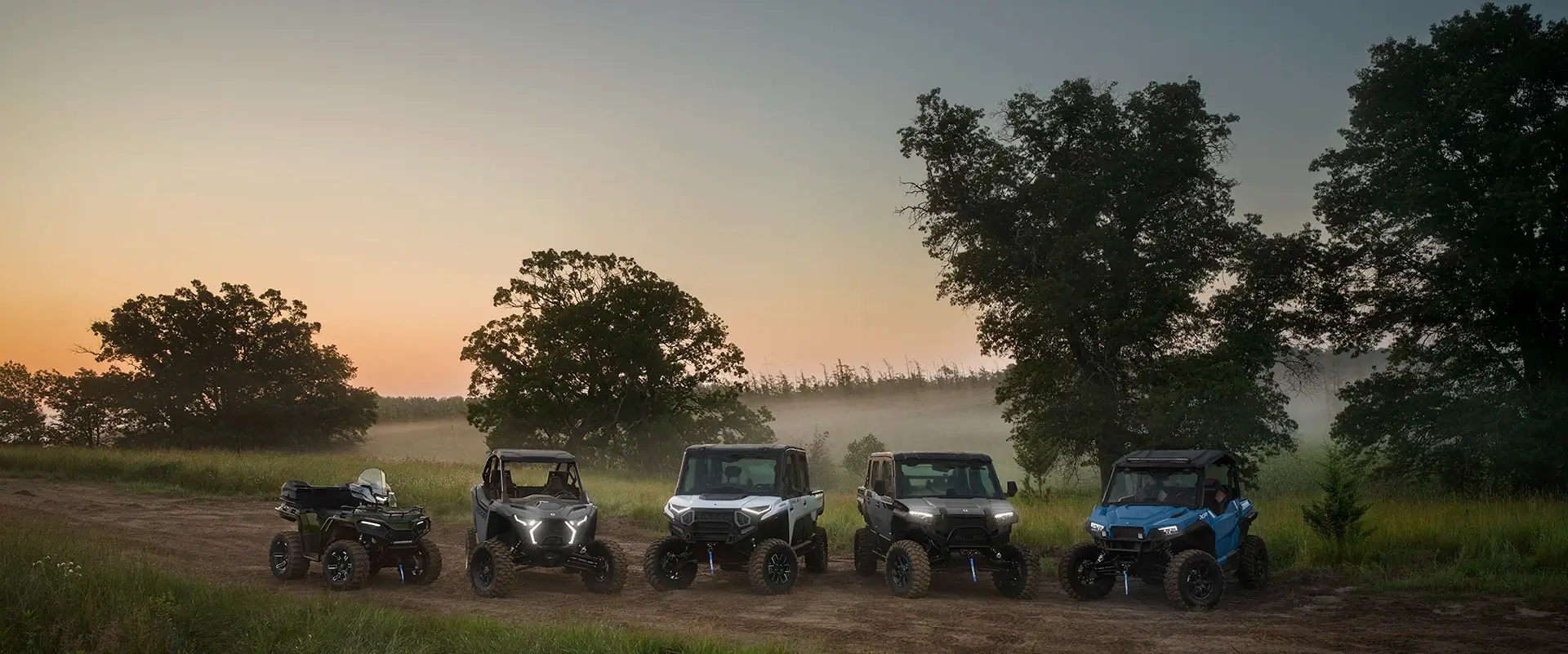 Polaris off-road vehicle lineup displayed on a rural trail at sunset featuring ATVs, side-by-side utility vehicles, and recreational models in an outdoor landscape