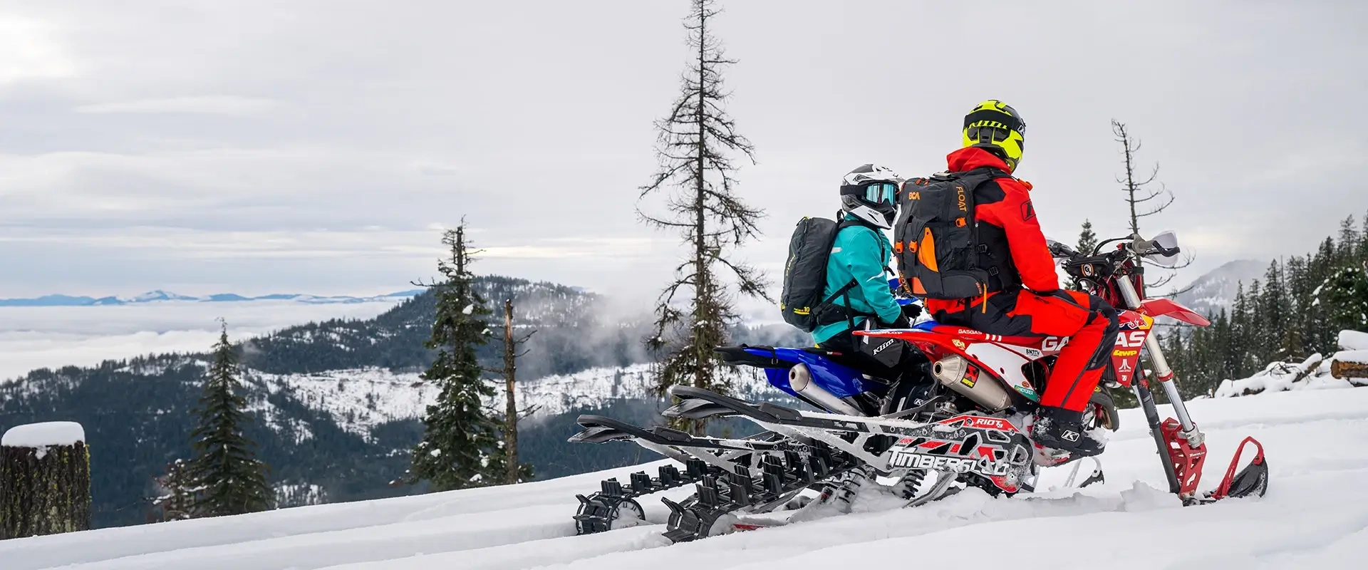 Two riders on red and blue Timbersled snow bikes parked on a snowy mountain ridge