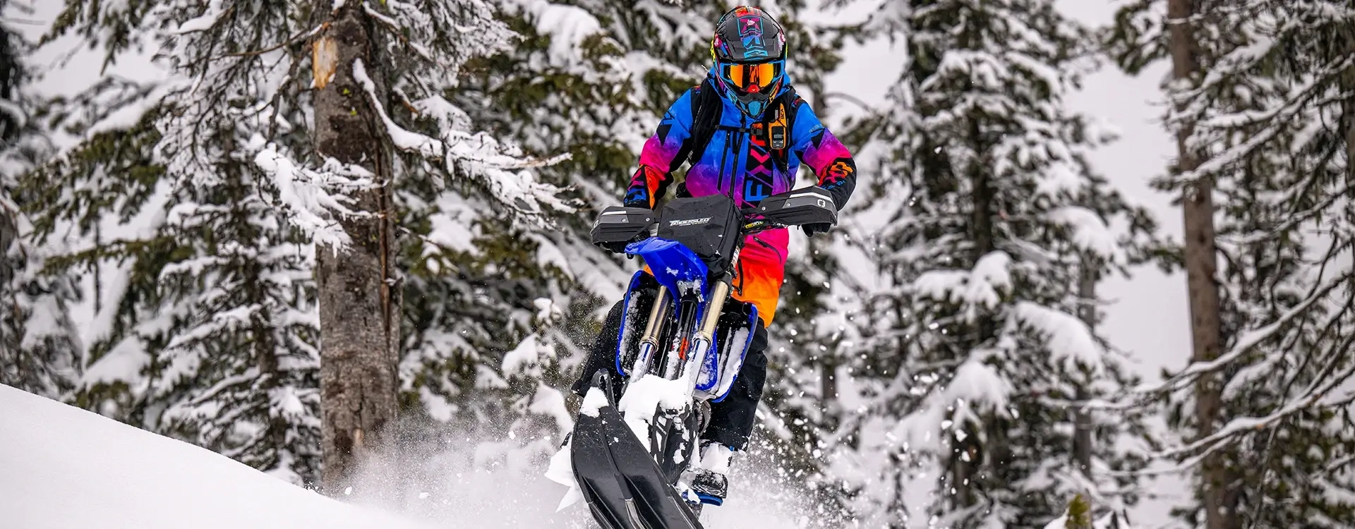 Person in bright colorful gear riding a blue Timbersled snow bike through a snowy forest